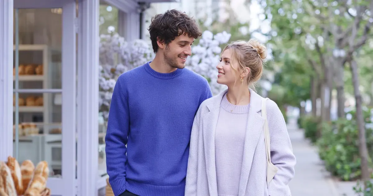 A young couple in their twenties smiling at each other on a quiet street outside a bakery.